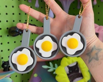 May include: Three gray plastic frying pan keychains, each featuring a raised white egg white and a bright yellow yolk in the center. Each pan has a small handle with a hole at the top, attached to a gold-toned metal ball chain for hanging.
