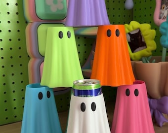 May include: A collection of six ghost-shaped beverage insulators in purple, lime green, orange, blue, white, and pink. Each ghost features two black oval eyes. One white ghost insulator holds a metal beverage can, while the others are empty.