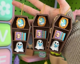 May include: Three brown coffin-shaped keychains featuring miniature shelves. Each shelf contains a small oval frame with a moon and clouds, a purple book with a skull on the cover next to blue books, and a white ghost figure holding a blue book.