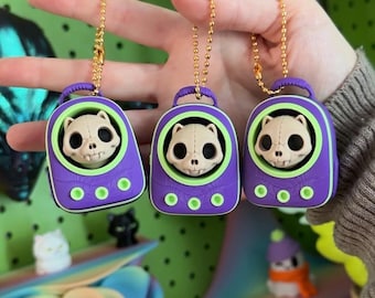 May include: Three identical purple and lime green keychain charms featuring a beige skull with large eye sockets inside a rounded housing. Each charm has a gold-toned ball chain attached to the top handle and three small green circular accents.