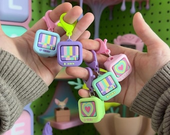 May include: A collection of five small television-shaped keychains in pastel colors including light blue, purple, pink, mint green, and lime green. Each keychain features a screen displaying either a colorful vertical stripe pattern or a heart.