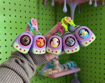 May include: Five small, portable keychain charms shaped like miniature purple and lime green pet carriers. Each carrier features a different animal head inside, including a brown dog, orange cat, yellow bird, gray cat, and blue bird, with plastic clips.