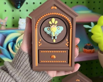 May include: A brown, house-shaped decorative object featuring a central circular clock-like face with a light blue and yellow moth emblem. The structure includes a rectangular drawer at the bottom with orange accents and small dot detailing.