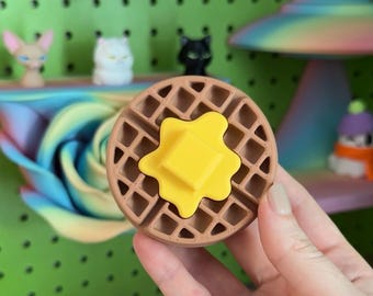 May include: A circular brown waffle-shaped object with a grid pattern and a bright yellow butter pat centered on top. The waffle has a textured surface and a realistic appearance, mimicking the look of a breakfast food item in a round shape.