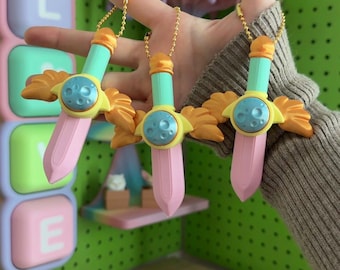 May include: Three sword-shaped keychains featuring a light blue handle, a pale pink blade, and a yellow wing-like crossguard. Each sword has a circular blue center piece with a moon-like texture and is attached to a gold-colored ball chain.
