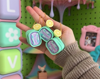 May include: Two miniature light blue sardine tin keychains featuring a purple label with the text SARDINES and a small yellow lemon slice charm attached to a gold-colored ball chain. The rectangular tins have rounded corners and a textured surface.