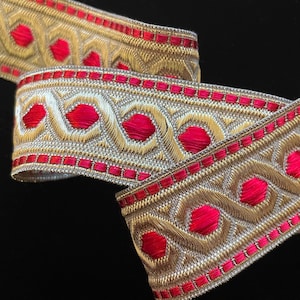 May include: Gold and red trim with a repeating pattern of circles and squares. The trim is woven and has a metallic sheen.