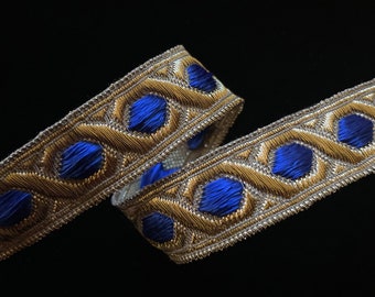 May include: Decorative ribbon with a gold and silver woven design, featuring blue circular accents. The ribbon is curled, showcasing its intricate pattern against a black background. Ideal for crafting and embellishments.