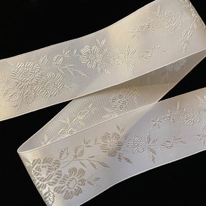 Puede incluir: Cinta de satén blanco con un diseño floral en relieve. La cinta tiene un brillo sutil y un delicado patrón floral.