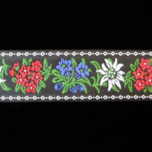 Puede incluir: Una cinta negra con borde blanco y un patrón repetido de flores rojas, azules y blancas. Las flores son estilizadas y se asemejan al edelweiss.