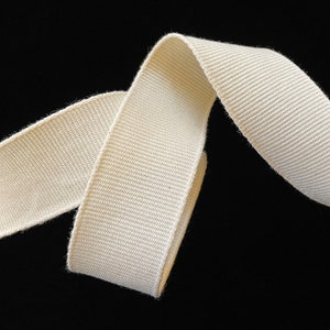 May include: Cream-colored woven ribbon against a black background. The ribbon is folded over itself, showing the texture of the fabric. The ribbon appears to be made of a sturdy material, suitable for various crafting projects.