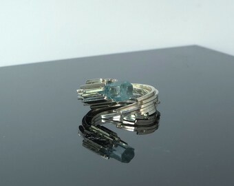 May include: A silver ring with a blue aquamarine gemstone set in the center. The ring is made of multiple thin bands of silver that are stacked and twisted together.