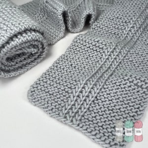 Peut inclure: Une écharpe en tricot gris avec un motif de point de câble. L'écharpe est enroulée et posée sur une surface blanche.