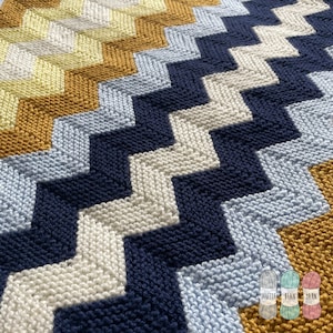 Könnte beinhalten: Eine gestrickte Babydecke mit Chevron-Muster in Gelb-, Blau- und Weißtönen. Die Decke ist aus weichem Garn gefertigt und perfekt zum Kuscheln.