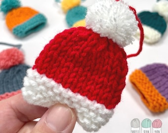 May include: A red and white knitted Santa hat with a white pom-pom. The hat is small and is being held in a hand. The hat is a handmade craft item.