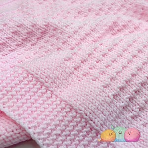 Puede incluir: Una manta tejida de color rosa claro con un patrón texturizado de rayas verticales alternas. La tela muestra una construcción consistente en punto bobo con crestas elevadas que crean un diseño geométrico repetitivo en la superficie.
