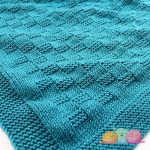 Könnte beinhalten: Eine türkisfarbene Strickdecke mit einem strukturierten, webartigen Muster. Die Decke ist aus weichem Garn gefertigt und weist ein sich wiederholendes quadratisches Design auf. Die Nahaufnahme hebt die detaillierten Nähte und das gemütliche Aussehen der Decke hervor.