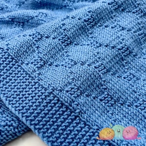 Puede incluir: Primer plano de una manta de punto azul claro. La manta presenta un patrón texturizado con cuadrados en relieve y un borde acanalado. El material suave y acogedor está doblado, mostrando el intrincado trabajo de puntadas y la profundidad del diseño.