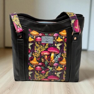May include: Black tote bag with a colorful mushroom print on the front panel. The bag has black handles and straps with a floral pattern. The text "SewFine by Lori" is visible on a small tag.