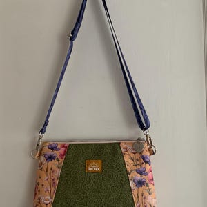 May include: A floral print crossbody bag with a green lace panel and a navy blue strap. The bag has a pink zipper and a small gold tag with the word "Sunshine" on it.