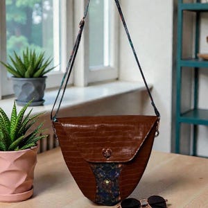 May include: A brown, crocodile-textured handbag with a patterned flap and strap. The bag has a heart-shaped clasp and a decorative panel. Round sunglasses rest on the wooden surface next to the bag.
