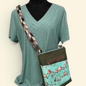May include: A crossbody bag featuring a dark olive green upper section and a light turquoise lower pocket decorated with a pattern of small red and blue birds perched on brown branches. The bag has an adjustable strap with a geometric chevron pattern.