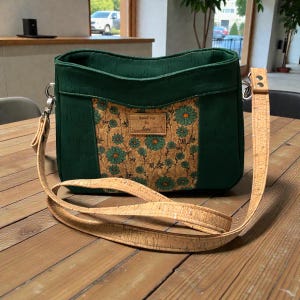 May include: Green crossbody bag with a cork floral design on the front. The bag has a long, adjustable cork strap and silver-tone hardware. The label on the front says "SewFine by tori".