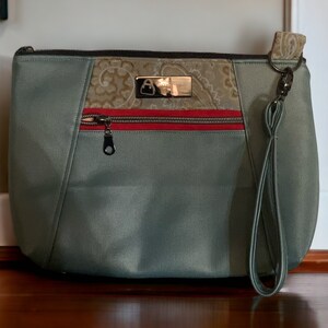 May include: A green zippered pouch with a brown and gold patterned fabric accent. The pouch has a red zipper and a gray strap.