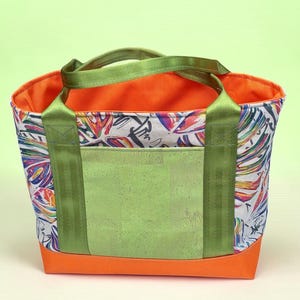 May include: A tote bag featuring a vibrant, multi-colored tropical leaf print on a white background. It has a solid orange base and interior lining, a light green textured front pocket, and olive green nylon webbing handles attached to the sides.