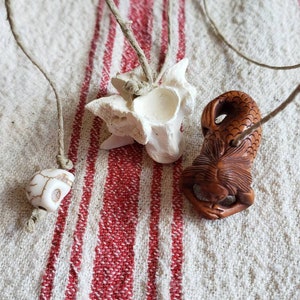 May include: Three unique pendant necklaces. One features a white and brown stone, another a bone-like structure, and the third a carved brown figure. Each is strung on a natural-colored cord, displayed on a red and white striped fabric.