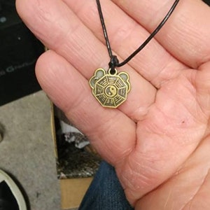 May include: A pendant necklace featuring a gold-colored charm with a Yin Yang symbol at the center. The charm is surrounded by an octagonal design. The necklace has a black cord.