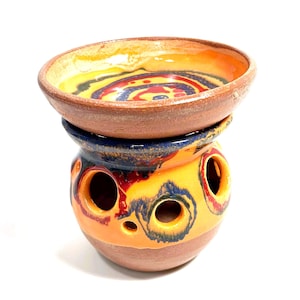 Pode incluir: Um aquecedor de óleo de cerâmica com uma base redonda com aberturas circulares e um prato superior removível. A peça tem um esmalte vibrante em tons de amarelo, laranja, vermelho e azul profundo, contrastando com a textura da argila castanha.