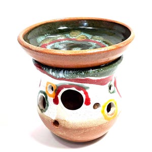 Może przedstawiać: Ceramiczny podgrzewacz do wosku z zaokrągloną podstawą pokrytą białą glazurą, ozdobiony kolorowymi czerwonymi, zielonymi i żółtymi wzorami kołowymi oraz kilkoma okrągłymi wycięciami. Na górze znajduje się pasująca płytka miseczka z ciemnozieloną i brązową glazurą.