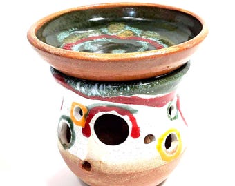 May include: A ceramic wax warmer featuring a rounded base with a white glaze, accented by colorful red, green, and yellow circular patterns and several circular cutouts. A matching shallow bowl with a dark green and brown glaze sits on top.