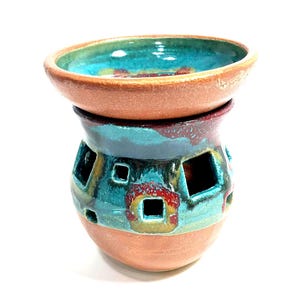 May include: A ceramic wax warmer featuring a wide, shallow bowl top and a rounded base with square cutouts. The piece displays a natural terracotta clay finish at the rim and bottom, contrasted by a vibrant teal, blue, and red glazed midsection.