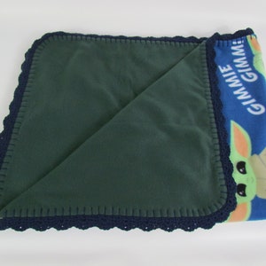May include: A green fleece baby blanket with a blue fleece side featuring a cartoon character with the text "Gimme Gimmie". The blanket has a navy blue crocheted border.