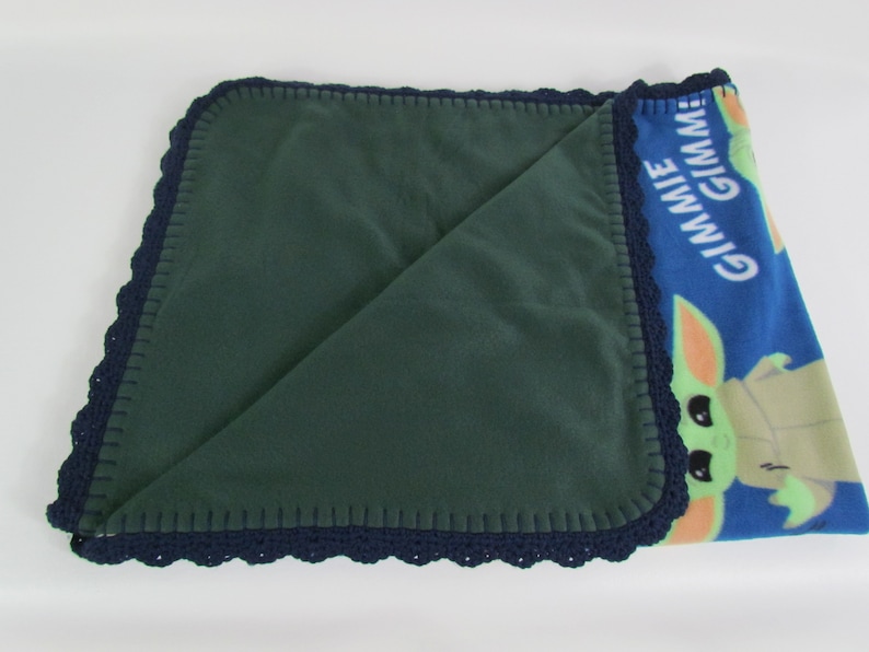 May include: A green fleece baby blanket with a blue fleece side featuring a cartoon character with the text "Gimme Gimmie". The blanket has a navy blue crocheted border.