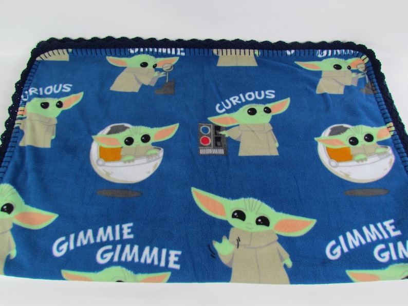 May include: Blue fleece blanket with a repeating pattern of a green alien character from the Star Wars series, The Mandalorian. The alien is wearing a brown robe and has large ears. The text "Curious" and "Gimme Gimme" is printed on the blanket.