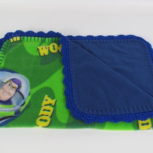 May include: A green and blue fleece blanket with a crocheted border. The blanket features a cartoon image of Buzz Lightyear from the Toy Story franchise. The blanket has the words "WOW" and "TODY" printed on it.