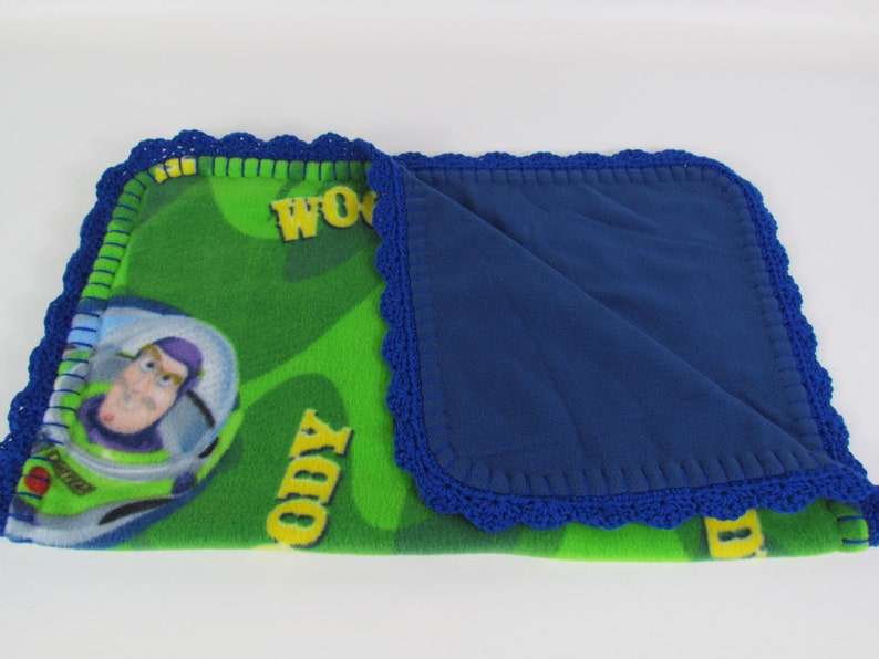 May include: A green and blue fleece blanket with a crocheted border. The blanket features a cartoon image of Buzz Lightyear from the Toy Story franchise. The blanket has the words "WOW" and "TODY" printed on it.