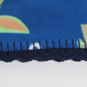 May include: Close-up of a blue fleece blanket with a crocheted navy blue border. The blanket has a colorful pattern with green, orange, and white accents.