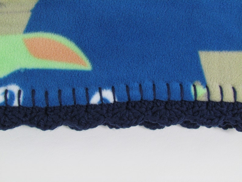 May include: Close-up of a blue fleece blanket with a crocheted navy blue border. The blanket has a colorful pattern with green, orange, and white accents.