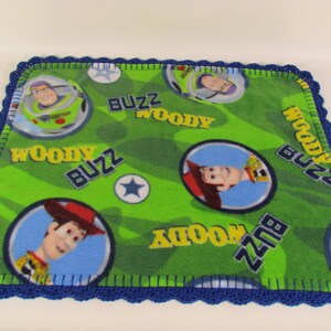 May include: Green fleece baby blanket with a blue crocheted border. The blanket features a repeating pattern of Woody and Buzz Lightyear from the Toy Story franchise. The words "Woody" and "Buzz" are repeated throughout the blanket.