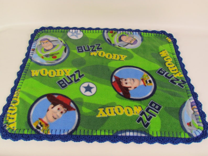 May include: Green fleece baby blanket with a blue crocheted border. The blanket features a repeating pattern of Woody and Buzz Lightyear from the Toy Story franchise. The words "Woody" and "Buzz" are repeated throughout the blanket.