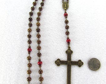 May include: A rosary featuring brown wooden beads, interspersed with smooth red beads, connected by metal links. It includes a central oval medal depicting a figure and a large, ornate, dark metal crucifix. A US quarter coin is placed for scale.