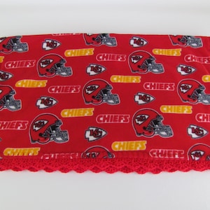 Pode incluir: Um cobertor de lã polar vermelha com um padrão repetido de capacetes de futebol americano, a palavra "CHIEFS" e o logotipo da equipa. O cobertor tem uma borda vermelha de crochê. Ideal para os fãs dos Chiefs.