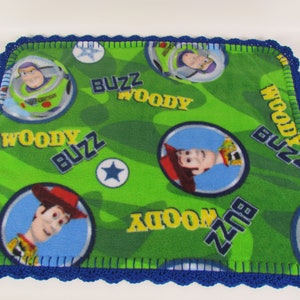 May include: A green fleece blanket with a blue crocheted border. The blanket features a repeating pattern of Woody and Buzz Lightyear from the Toy Story franchise. The blanket has the words "Woody" and "Buzz" printed on it.