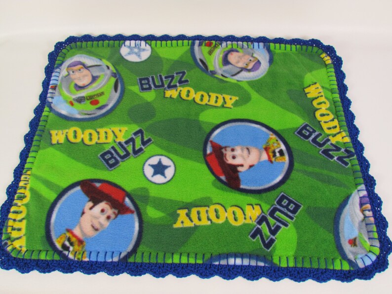 May include: A green fleece blanket with a blue crocheted border. The blanket features a repeating pattern of Woody and Buzz Lightyear from the Toy Story franchise. The blanket has the words "Woody" and "Buzz" printed on it.