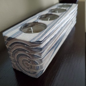 May include: A wooden candle holder with a natural wood grain finish and three circular candle wells. The holder is made of stacked wood planks with a dark grey stain.