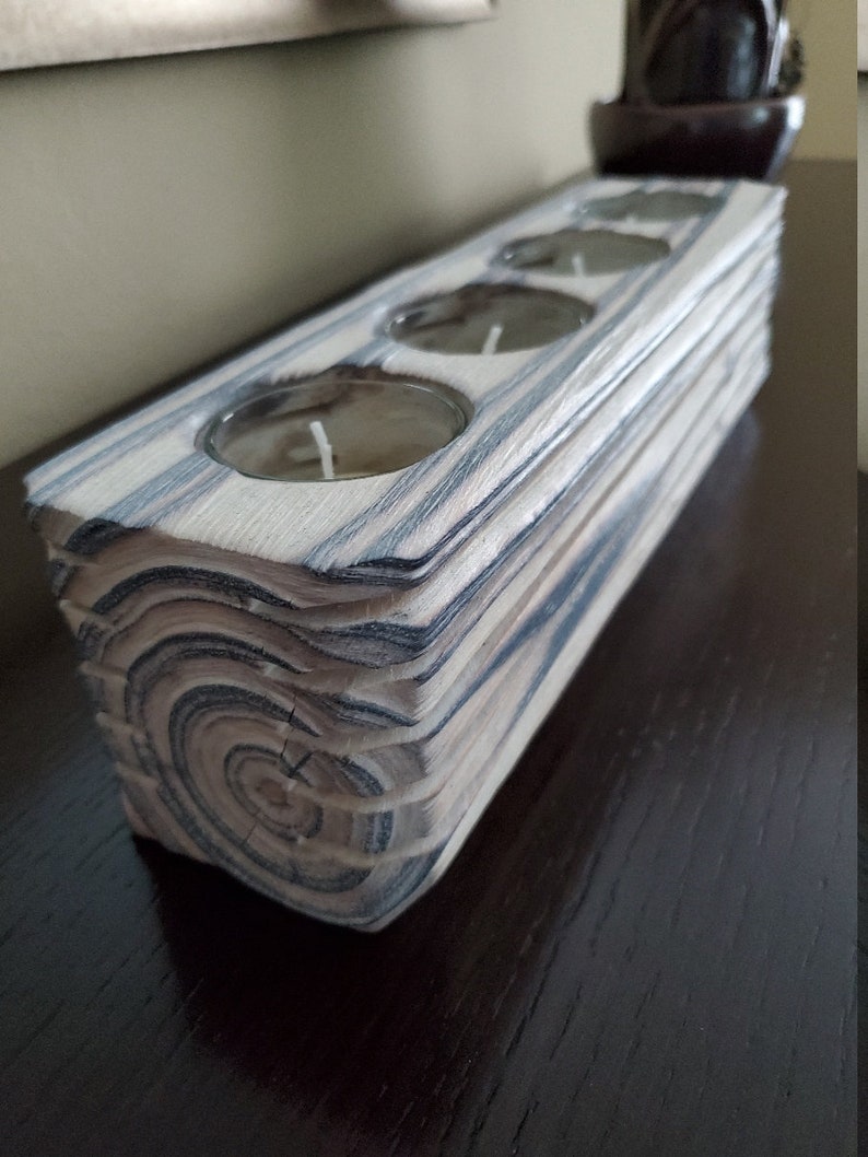 May include: A wooden candle holder with a natural wood grain finish and three circular candle wells. The holder is made of stacked wood planks with a dark grey stain.